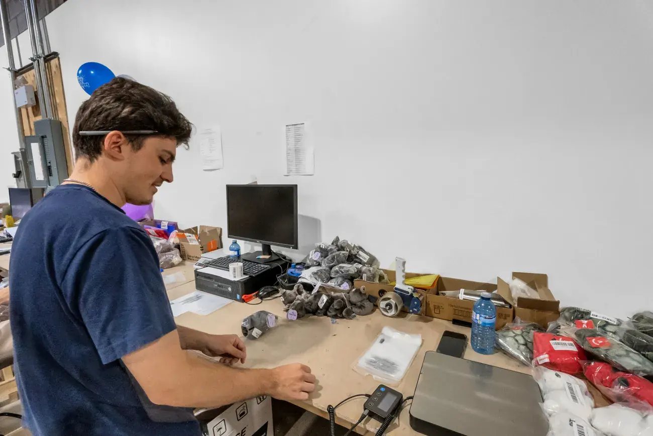 A Prep Buddies team member prepping stuffed toys at a wooden bench with a label printer, tape gun and shipping scale