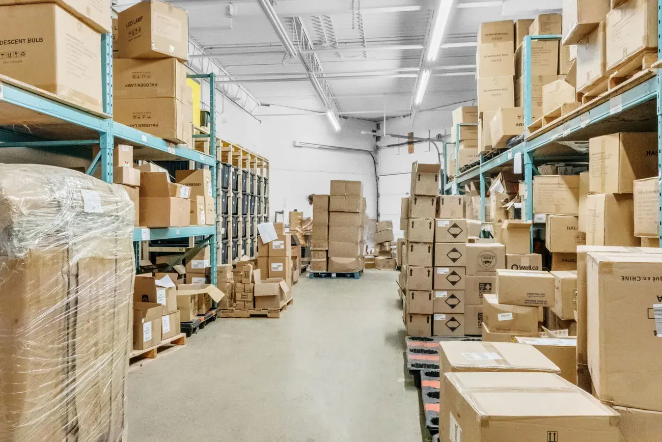 A narrow aisle between teal pallet racking stacked with cartons, with black storage bins in wooden racks at the far end
